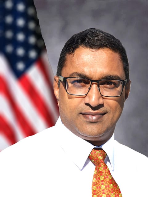 Suresh Vuyyuru, in a white shirt and patterned tie, standing beside a United States flag.